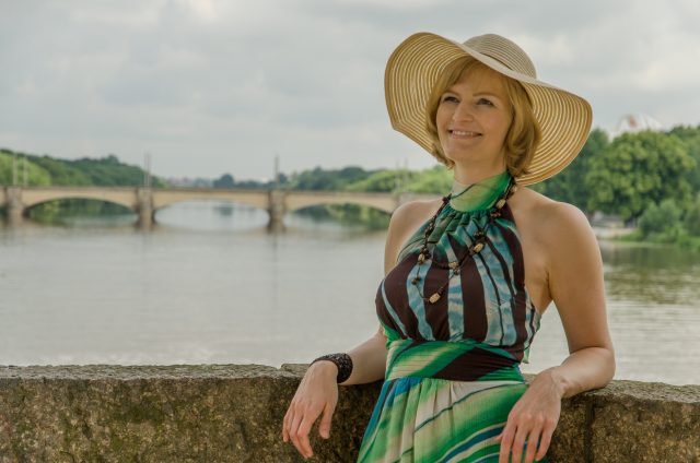 Lebensfrohe, junge Frau mit Hut und Sommerkleid auf einer Brücke mit Fluss, Brücke und Grün im Hintergrund